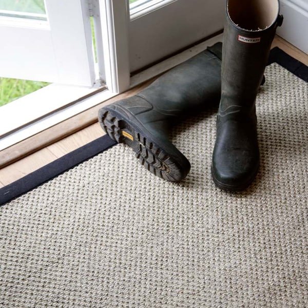 A beige rug with a black border placed by a door with rubber boots on top.