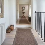 A bright, rustic hallway featuring two long sisal runner rugs on light wood plank floors, leading toward a doorway with a small console table.