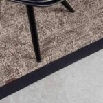A high-angle, close-up shot of a textured brown and beige woven rug with a thick black border, featuring a black chair leg resting on it.