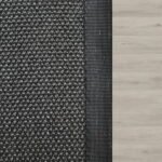 Top-down view of a dark grey textured rug with a black border on light wood flooring.